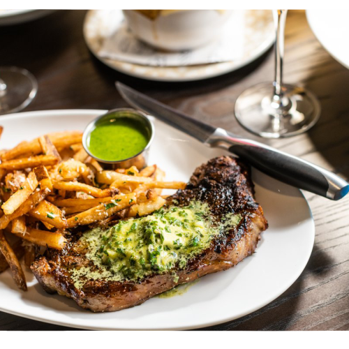 A plated grilled steak with green herb sauce, a side of fries, and a blue drink on a dining table.
