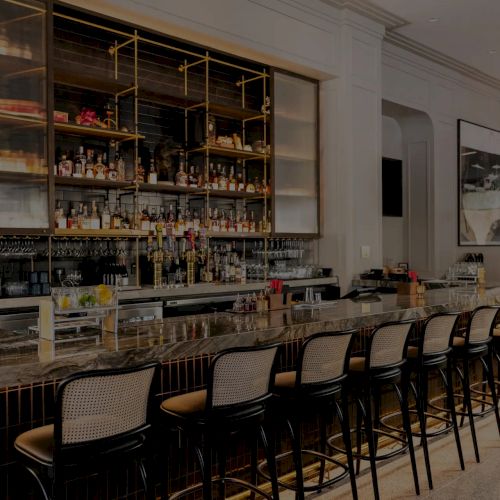 A stylish bar interior with open shelves of liquor, glassware, and stools lined up along a marble counter.