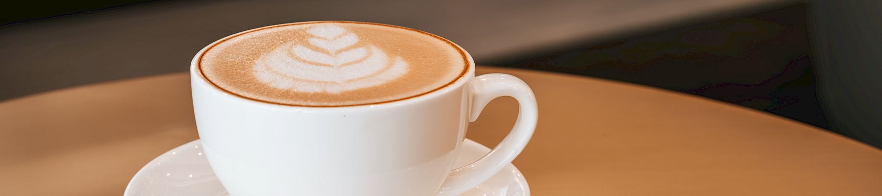 A white cup of cappuccino/latte with warm latte art on top, served on a saucer in a cozy caf&eacute; setting.