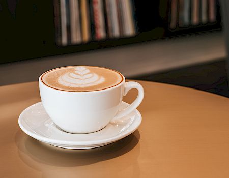 A white cup of cappuccino/latte with warm latte art on top, served on a saucer in a cozy café setting.